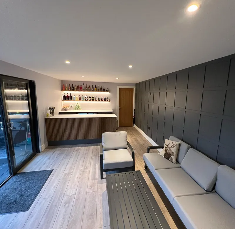 Finished garden room with a dark panelled feature wall, lit back-bar shelving and outdoor sofa